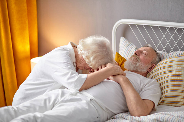 Senior woman sitting next to dying husband lying on bed and crying - Foto, imagen