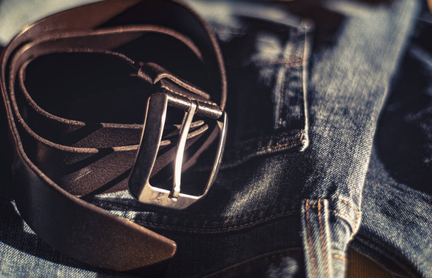 leather belt is a casual wardrobe accessory. Beautiful leather belt and jeans close-up. Men's brutal accessories, belt on a dark background with blurred depth of field, brown belt and denim - Photo, Image