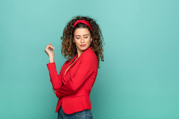 Positive self assured female model with long curly hairstyle wearing stylish bright red jacket and headband standing against green background - Photo, Image