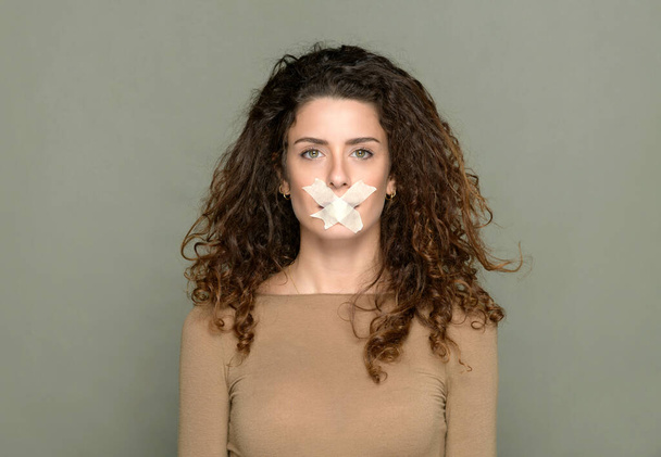 Speechless female with cross shaped tape covering mouth on gray background in studio and looking at camera while showing silence concept - Photo, Image