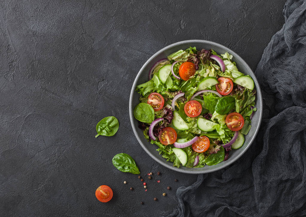 Fresh healthy vegetarian vegetables salad with tomatoes and cucumber, red onion and spinach in grey bowl plate on dark background with black kitchen cloth.Top view. - Photo, Image