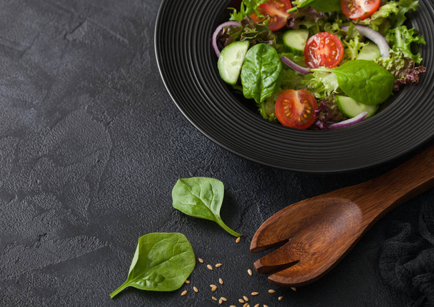 Healthy fresh vegetables salad with lettuce and tomatoes, red onion and spinach in black bowl on dark table background with spatula spoon - Photo, Image