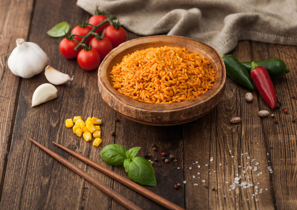 Wooden bowl with boiled red long grain basmati rice with vegetables on wooden table background with sticks and tomatoes with corn,garlic and basil with hot pepper. - Foto, Imagen