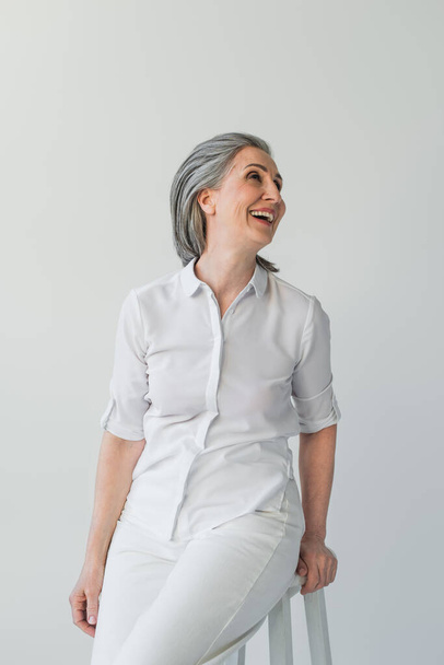 Grey haired woman in white shirt looking away on chair isolated on grey  - Foto, afbeelding