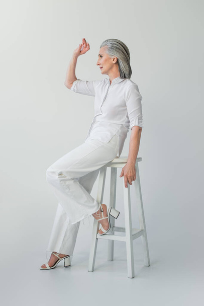 Mature woman in white clothes looking away on chair on grey background  - Foto, immagini