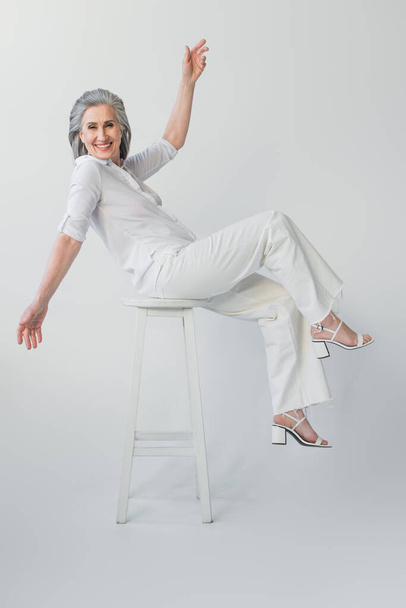 Happy mature woman looking at camera on chair on grey background  - Foto, Imagem