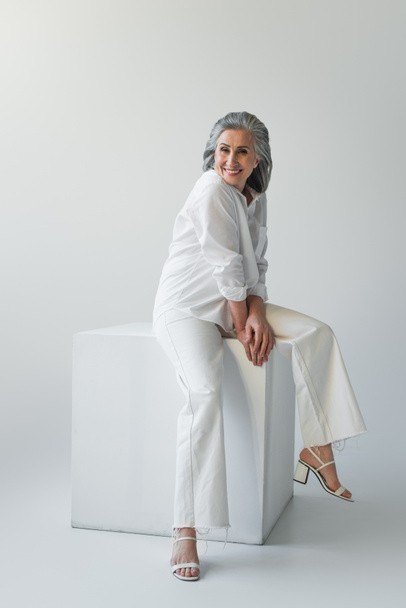 Smiling mature woman sitting on white cube on grey background  - Foto, afbeelding