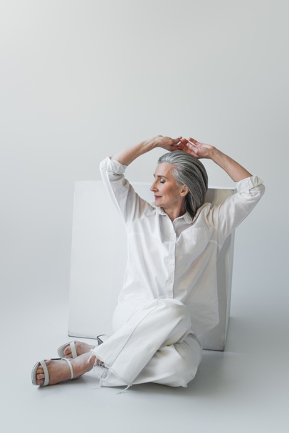 Grey haired woman posing near white cube on grey background  - Фото, изображение