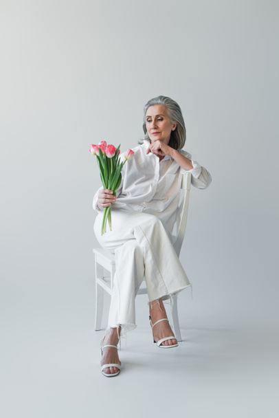 Mature woman looking at flowers while sitting on chair on grey background  - Photo, Image