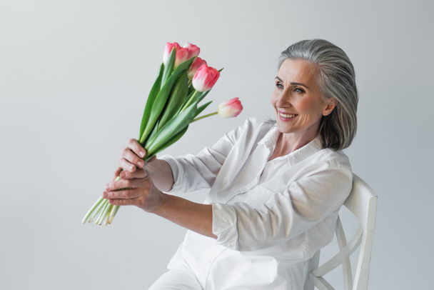 Ältere Frau lächelt beim Betrachten von Tulpen auf Stuhl isoliert auf grau  - Foto, Bild