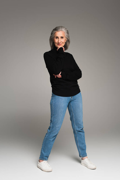 Full length of mature woman in jeans and turtleneck looking at camera on grey background  - Foto, afbeelding
