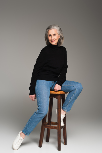 Mature woman sitting on chair and smiling on grey background  - Fotografie, Obrázek