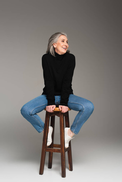 Positive woman in jeans and jumper sitting on chair on grey background  - Foto, imagen