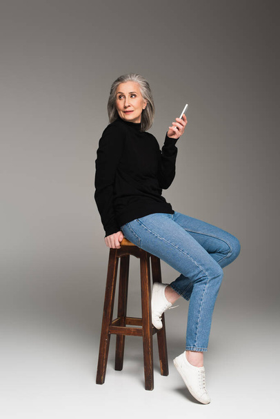 Mature woman in jeans holding smartphone on chair on grey background  - Photo, Image