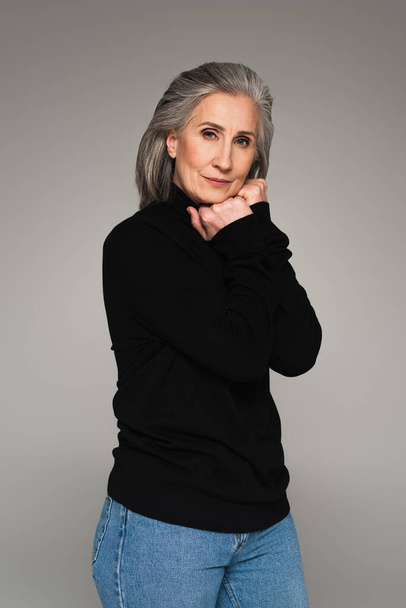 Mature woman in black jumper and jeans looking at camera isolated on grey  - 写真・画像