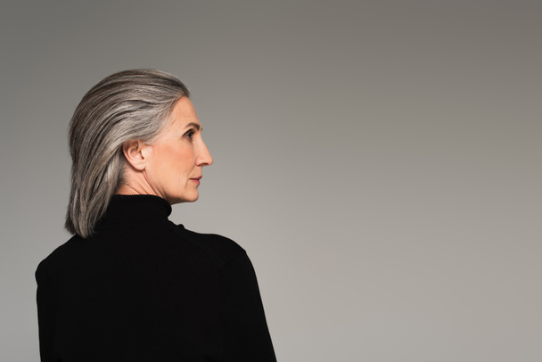 Vista lateral de mujer madura en cuello alto mirando hacia otro lado aislado en gris - Foto, imagen