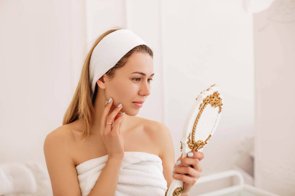 Een jonge vrouw in een witte handdoek kijkt naar zichzelf in de spiegel na de ingreep bij de schoonheidsspecialiste. De vrouw onderzoekt haar gezicht. Huidverzorging.  - Foto, afbeelding