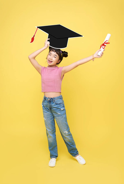 Happy Asian cute schoolgirl with graduation hat and diploma isolated on yellow background. - Zdjęcie, obraz