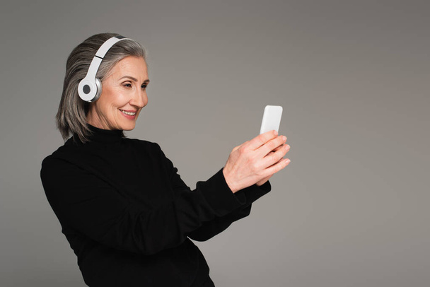 Usmívající se starší žena ve sluchátkách pomocí smartphone izolované na šedé  - Fotografie, Obrázek