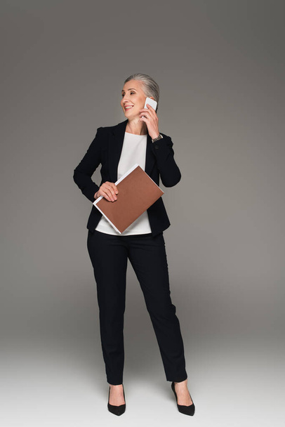 Smiling businesswoman with paper folder talking on smartphone on grey background  - Valokuva, kuva
