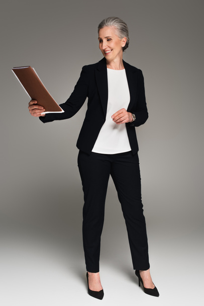 Улыбающаяся взрослая деловая женщина смотрит на бумажную папку на сером фоне  - Фото, изображение