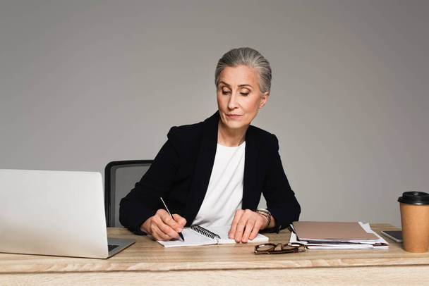 Donna d'affari che scrive su taccuino vicino a gadget e carte isolate su grigio  - Foto, immagini