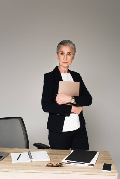 Businesswoman holding paper folder near notebook and eyeglasses on table isolated on grey  - Φωτογραφία, εικόνα