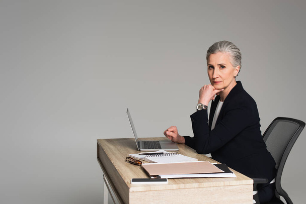 Mature businesswoman looking at camera near gadgets and papers on table isolated on grey  - Foto, Bild