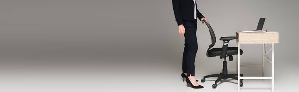 Cropped view of businesswoman standing near office chair and laptop on working table on grey background, banner  - Photo, Image