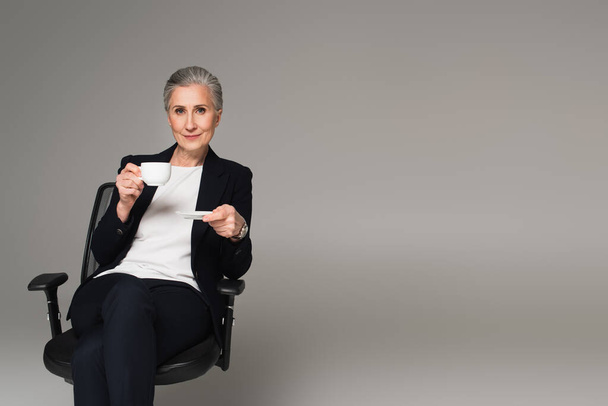 Mature businesswoman holding cup of coffee on office chair isolated on grey  - Photo, image