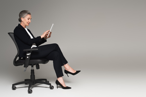 Mature businesswoman using digital tablet on office chair on grey background with copy space  - Φωτογραφία, εικόνα