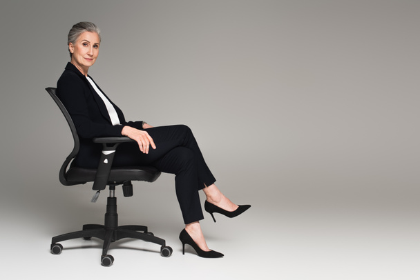 Middle aged businesswoman with crossed legs looking at camera on office chair on grey background  - 写真・画像