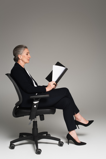 Side view of mature businesswoman looking at documents on office chair on grey background  - Foto, Imagen