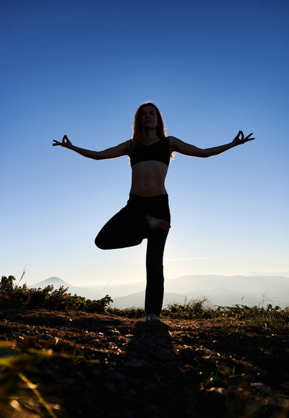 Full length of young woman performing yoga pose on grassy hill with blue sky on background. Female person standing on one leg and doing Gyan mudra hand gesture while practicing yoga outdoors. - Photo, Image