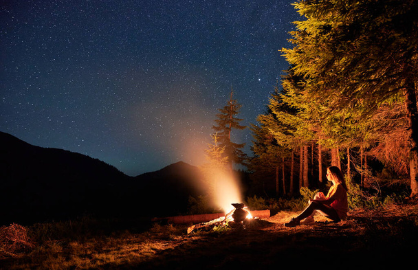 キャンプファイヤーの近くに木や女性旅行者と芝生の丘の上の夜空の壮大な景色。若いです女性ハイカー座って近くたき火の下で美しいです空とともに星. - 写真・画像