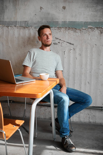 Férfi ül az asztalnál laptoppal és egy csésze kávéval. A fiatalember munka után pihen a neten. A hazai iroda fogalma. - Fotó, kép