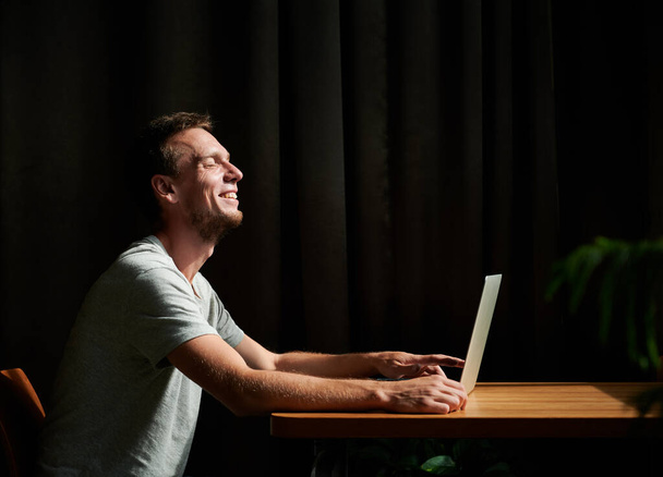 Vista lateral de hombre sonriente estudiante sentado junto a la mesa y trabajando en el ordenador portátil en casa. Concepto de oficina en casa. - Foto, Imagen
