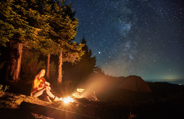 Jovem mulher relaxando na natureza sob céu estrelado perto da fogueira. Conceito de acampamento de verão noturno em montanhas. - Foto, Imagem