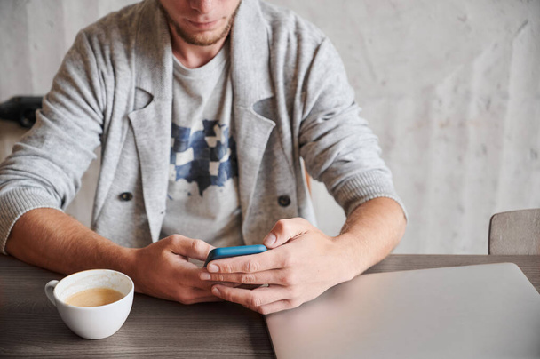 Cropped view of young man with beard sitting on chair with smartphone and cup coffee and modern laptop. Concept of studying or working in phone at home. - Photo, Image