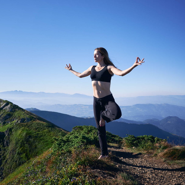 Полная длина спортивной женщины в спортивной одежде, балансирующей на одной ноге и делающей жест руки Gyan mudra во время занятий йогой на открытом воздухе. Молодая спортсменка, занимающаяся йогой в горах. - Фото, изображение