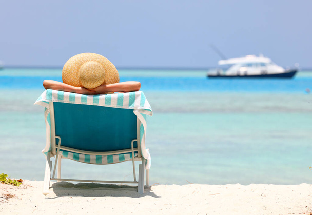 Visão traseira de uma fêmea tomando banho de sol em um chapéu em uma espreguiçadeira durante umas férias de verão na praia junto ao oceano - Foto, Imagem