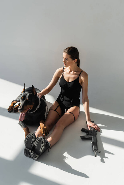 high angle view of woman in black bodysuit sitting near rifle and doberman on grey background with shadows - Photo, Image