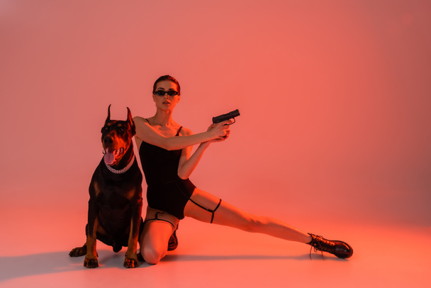 young woman in black bodysuit holding handgun near doberman on pink background with yellow light - Foto, afbeelding