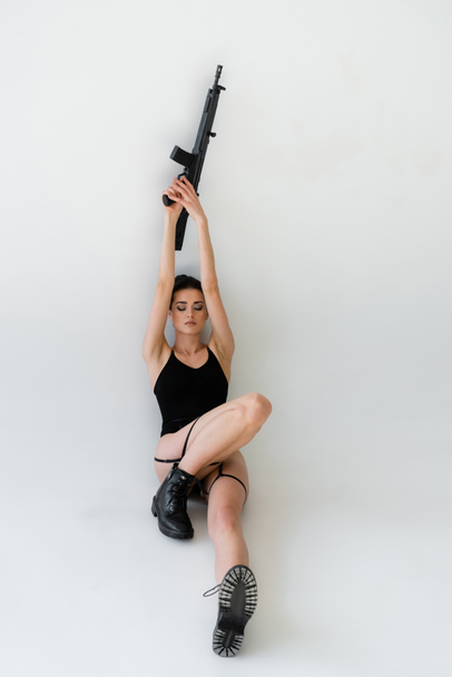 sensual woman holding rifle in raised hands while sitting with closed eyes on grey background - 写真・画像
