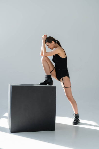 stijlvolle vrouw in bodysuit stepping op zwarte kubus op grijze achtergrond met schaduwen - Foto, afbeelding