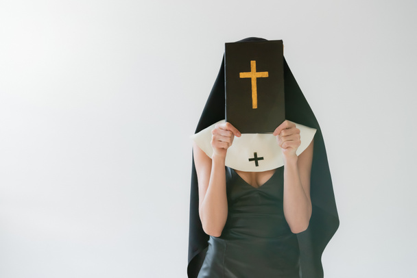 nun in sexy dress obscuring face with holy bible isolated on grey - Photo, Image
