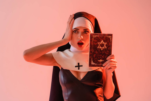 σοκαρισμένη καλόγρια σε σέξι φόρεμα αγγίζοντας το κεφάλι, ενώ κρατώντας εβραϊκή Βίβλο απομονωμένη σε ροζ - Φωτογραφία, εικόνα