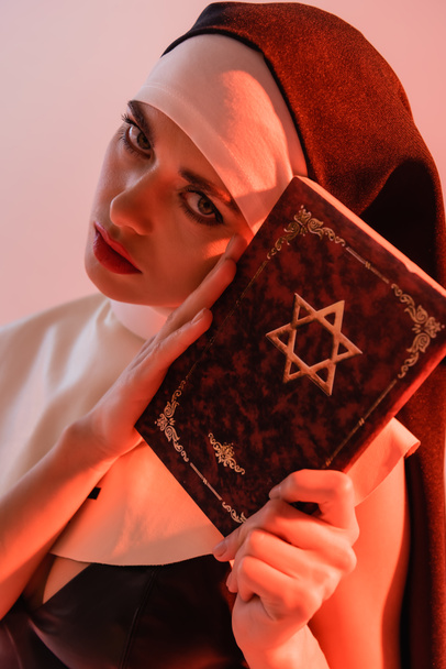 jovem, freira sexy olhando para a câmera enquanto segurando bíblia judaica isolado em rosa - Foto, Imagem