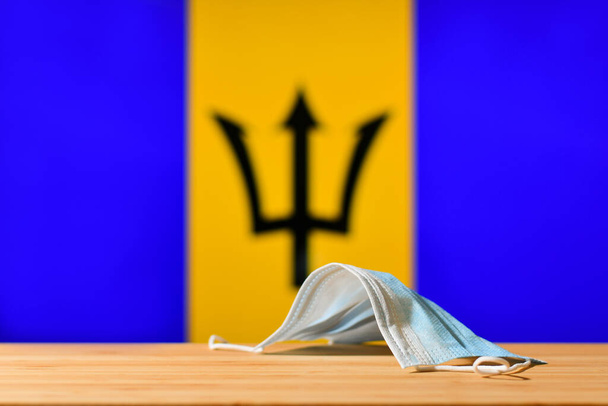 Медицинская маска лежит на столе на фоне флага Барбадоса. Концепция обязательного режима маски для жителей страны и туристов на Барбадосе во время пандемии. - Фото, изображение