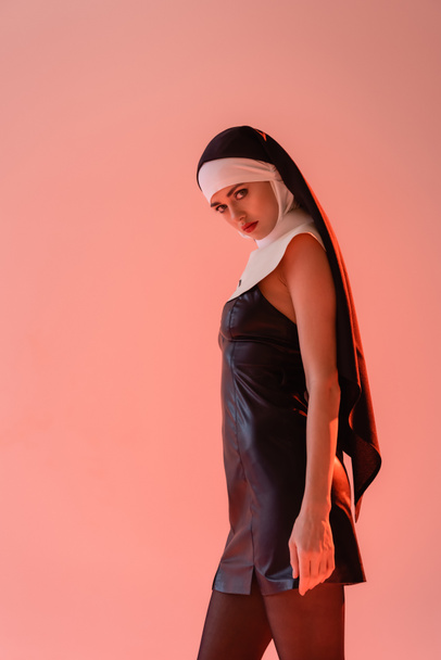 young nun in sexy dress looking at camera while standing isolated on pink - 写真・画像